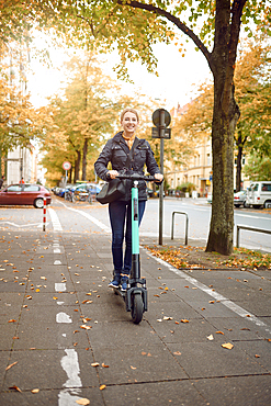 Young happy blond woman riding an electric scooter in the city, smiling at the camera, in autumn