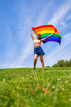 Caucasian brunette woman with a rainbow lgbt flag on the grass and blue sky background, waving the flag