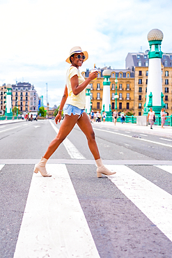 Portrait of black ethnic woman drinking an orange juice enjoying summer on vacation crossing the street at the zebra crossing