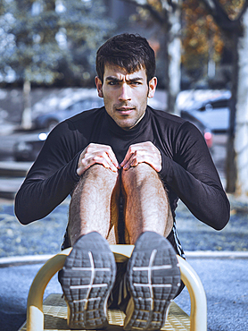 Front view of a young man doing abdominal exercises on a public equipment in outdoor fitness