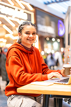 Alternative girl with white braids with a computer in a shopping center, looking at the camera with a hot coffee in her hands