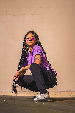 Urban session. Dark-skinned young woman with long braids and purple glasses, on the floor with a smooth wall in the background