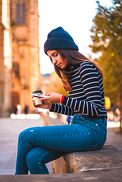 Lifestyle, brunette Caucasian sending a message with her mobile phone in the city. Relaxing with a take away coffee next to a beautiful church in the park. Black sweater and with a wool hat