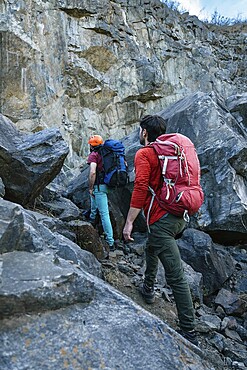 Two hikers with backpacks approach a rocky cliff, preparing for a challenging climb. They are wearing helmets and climbing gear, ready for an outdoor adventure