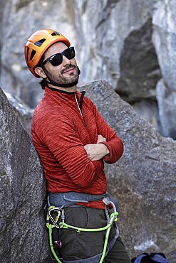 A confident sunglassed climber in orange helmet and red jacket rests against a rock. He smiles with his arms crossed, showing off his climbing gear in a rugged outdoor setting