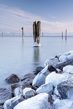 A wooden pile and ice-covered stones on a calm body of water under a blue sky in winter, harbour, Immenstaad, Lake Constance, Baden-Württemberg, Germany, Europe