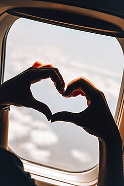 Female hand in heart shape Airplane flight. View from the window of the plane. Airplane, Aircraft. Vacation love travel concept Traveling by air. Airplane Window View Above the Clouds Amazing golden fluffy clouds moving softly on the sky aerial view