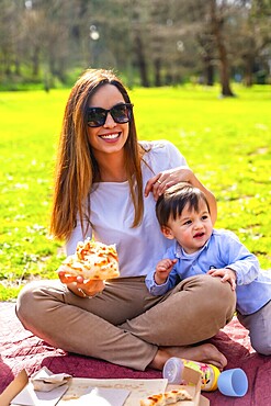 Happy mother sharing a slice of pizza with her son during a sunny picnic in the park, enjoying quality time and laughter together