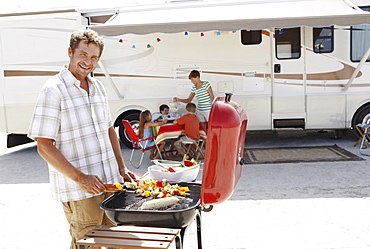 Man barbecuing with family and motor home in background