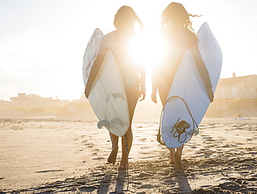 Two female surfers walking on beach at sunset, Rockaway Beach, New York