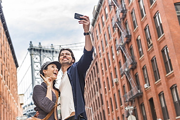 Couple taking selfie on street, Brooklyn Bridge in background, Brooklyn, New York