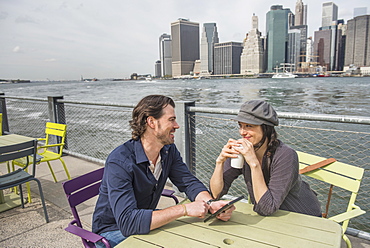 Happy couple sitting and using tablet pc and drinking coffee with cityscape in background, Brooklyn, New York