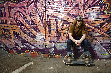 Young man with skateboard in front of graffitied wall