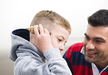 Father watching son hold seashell up to ear