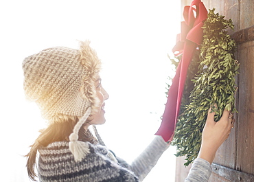 Side view of young woman hanging Christmas wreath on entrance door