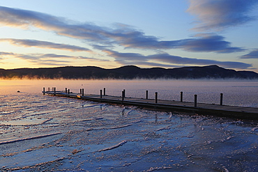 Jetty on frozen lake with vapor, hills in background at sunrise, Lake George, New York