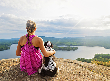 Young woman sitting with dog on cliff looking at lake view, USA, Maine, Camden