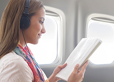 Young woman wearing headphones while reading book on plane