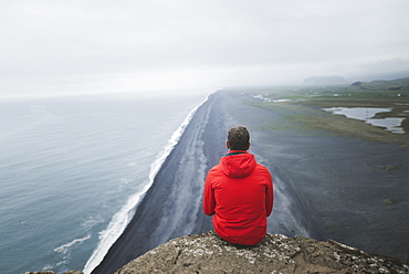 Man wearing red coat sitting above beach in Vik, Iceland