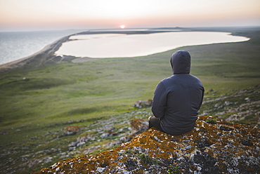 Young man in hooded jacket sitting on hill during sunset