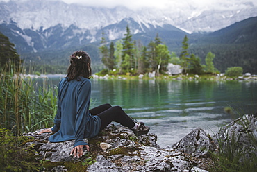 Germany, Bavaria, Eibsee, Young woman sitting on rock at shore of Eibsee lake in Bavarian Alps