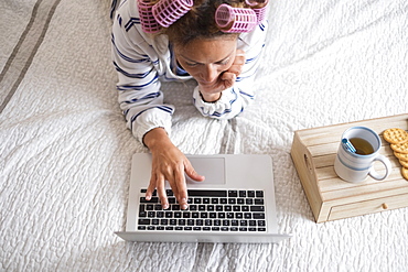 Woman with hair curlers lying on bed and using laptop