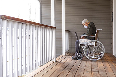 Senior man in wheelchair wearing protective mask to prevent coronavirus transmission using laptop on porch