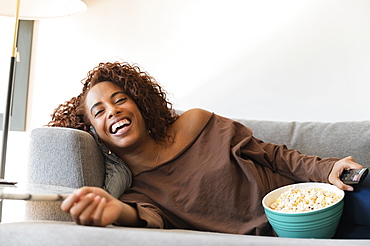 Laughing woman on sofa with TV remote and popcorn