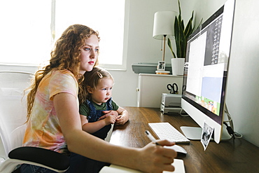 Young mother with her daughter on lap working from home at desk