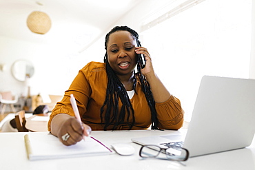 Woman working at desk at home, writing and talking on mobile phone