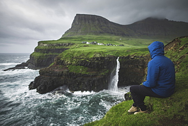 Denmark, Faroe Islands, Gasadalur Village, Mulafossur Waterfall, Man sitting on edge of cliff and looking at Mulafossur Waterfall