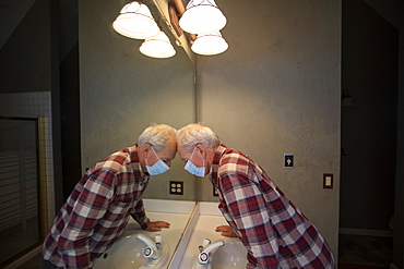 Senior man in Covid protective mask leaning head on mirror in bathroom