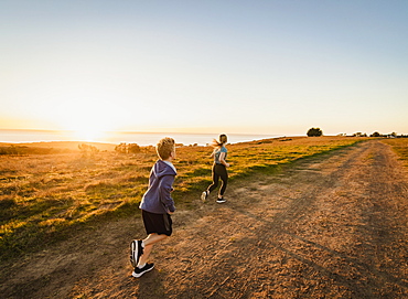 United States, California, Cambria, Rear view of boy (10-11) and girl (12-13) running on footpath in landscape at sunset