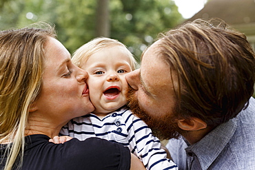 Mother and father kissing baby girl on cheek, outdoors