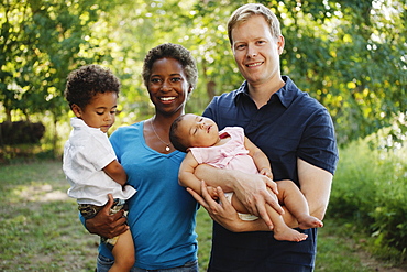 Family outdoors posing for photograph holding preschool boy and baby girl looking at camera smiling