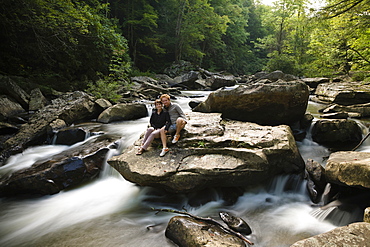 Portrait of mid adult couple, sitting together on rock in waterfall, New River Gorge National River, Fayetteville, West Virginia, USA