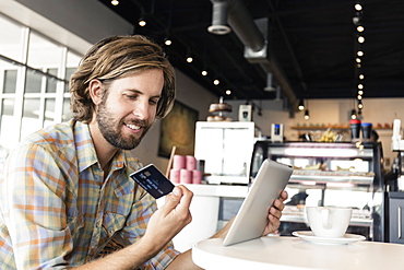 Mid adult man in coffee shop, using digital tablet, holding credit card