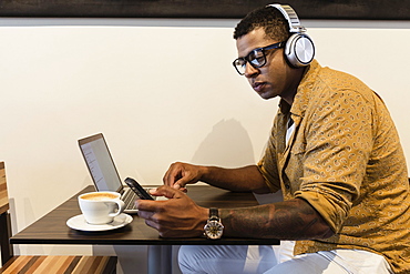 Young man in coffee shop, wearing headphones, using laptop and smartphone