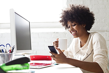 Young woman at desk with coffee using smartphone