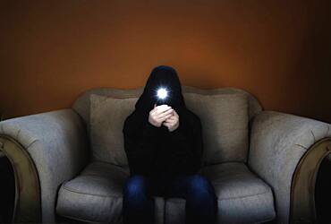 Man in hood sitting on sofa and holding flashlight in front of his face