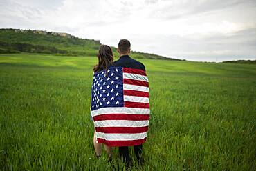 Rear view of young couple wrapped in American flag standing in wheat field