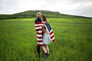 Smiling young couple wrapped in American flag in wheat field