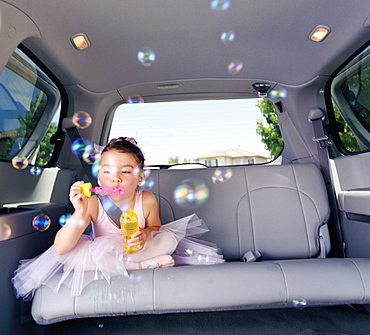 Girl (6-7) in ballet outfit blowing bubbles in car