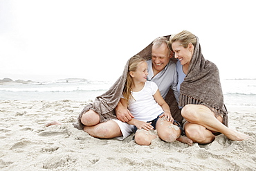 Smiling parents with daughter (10-11) on coastline covered by blanket