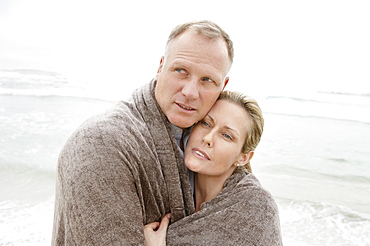 Embraced couple covered by blanket looking away on beach