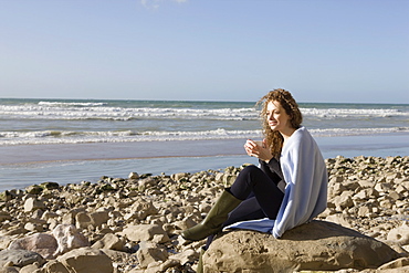 France, Pas-de-Calais, Escalles, Woman wrapped in blanket sitting on rocky beach, France, Pas-de-Calais, Escalles