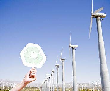 USA, California, Palm Springs, Coachella Valley, San Gorgonio Pass, Woman's hand holding recycling sign against blue sky and wind turbines