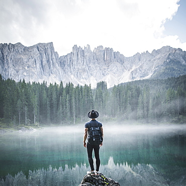 Italy, Carezza, Young man standing on rock at Lago di Carezza in Dolomite Alps at dawn