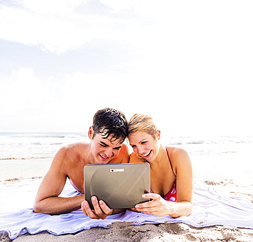 Portrait of young couple lying on blanket on beach side by side, using digital tablet, Jupiter, Florida