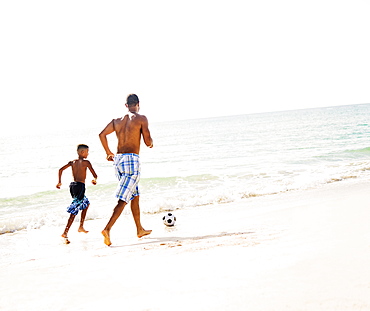 Father and son (10-11) playing soccer on beach